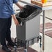 A person putting a black garbage bag into a Lavex grey rectangular under-counter trash can.