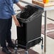 A woman's hand putting a black garbage bag into a Lavex black rectangular under-counter trash can.