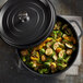 A black non-stick round casserole dish with a cover, filled with roasted vegetables.