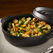 A black non-stick round casserole dish with a cover, filled with roasted vegetables.