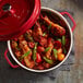 A red non-stick round casserole dish with a cover, filled with cooked chicken drumsticks, vegetables, and herbs.