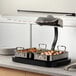 A stainless steel roasting pan with handles, shown under a food warming lamp on a countertop.