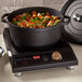 A black non-stick round casserole dish filled with cooked food, placed on an induction cooktop with a digital display and a control knob.