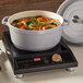 A gray non-stick round casserole dish with handles and a matching lid, shown on an induction cooktop and filled with soup.