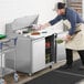 A man in an apron and gloves using an Avantco stainless steel sandwich prep table to store a plastic container of vegetables.