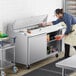 An Avantco stainless steel sandwich prep table with a man in an apron preparing food.