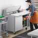 An Avantco stainless steel sandwich prep table with a man in an apron preparing food.