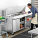 A man in an apron using an Avantco sandwich prep table to fill a container with food.