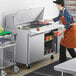 A woman in an apron using an Avantco stainless steel sandwich prep table to put food into containers.