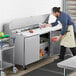 An apron-wearing man using an Avantco sandwich prep table to hold containers of food.