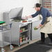 A man in an apron is preparing food in a kitchen using an Avantco stainless steel sandwich prep table with a cutting board.