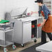 An aproned man in a commercial kitchen using an Avantco stainless steel sandwich prep table to put food into a container.