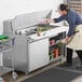 A man in a white apron using an Avantco ADA height refrigerated sandwich prep table to prepare food.