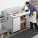 An aproned man in a school kitchen using an Avantco stainless steel sandwich prep table to put food in a container.