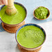A hand using a bamboo whisk to mix Jade Leaf Ceremonial Matcha in a bowl of green liquid.