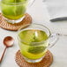 A glass cup of Jade Leaf Organic Matcha green tea with a tea bag in it and a spoon.