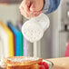 A person pouring coarsely ground cheese from a clear polycarbonate shaker onto a plate of food.