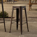Two Lancaster Table & Seating copper outdoor barstools with tan cushions on a stone patio.