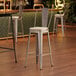 A Lancaster Table & Seating silver barstool with a tan cushion on a wood floor.