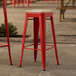 A red Lancaster Table & Seating outdoor barstool with a tan cushion on a stone surface.