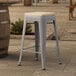 A Lancaster Table & Seating gray metal outdoor counter stool with a tan cushion on a stone surface.