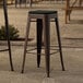 A pair of Lancaster Table & Seating copper outdoor bar stools with black fabric cushions on a stone surface.