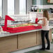 A red buffet or salad bar with a free-standing sneeze guard, featuring various food trays and a person serving themselves.