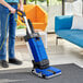 A person using a Clarke walk behind floor scrubber with a yellow cord to clean a rug in a room.