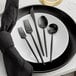 A white plate with a black Acopa Odin flatware set consisting of a knife, fork, and spoon.