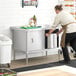 A man in an apron pushing a Steelton stainless steel enclosed base table with sliding doors.