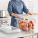 A woman in a chef's uniform holding a Vigor clear polycarbonate food storage container full of apples.