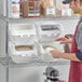 A man in an apron using a Baker's Lane ingredient shelf bin to scoop rice into a white container with a clear lid.