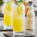 A close-up of a glass of yellow liquid with a lime slice on the rim with a bottle of Fee Brothers Toasted Almond Bitters.