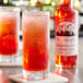 A glass of red liquid next to a bottle of Fee Brothers Maraschino Cherry Syrup.