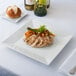 A 14" Bright white square porcelain plate with chicken and vegetables on a table.