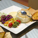 A 14" bright white square porcelain plate with food and a spoon on a table.