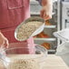 A person holding a Choice aluminum scoop of oats over a clear bowl.