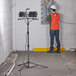 A man wearing a white helmet and orange safety vest standing in a room with two Genesis dual-head LED work lights on a stand.