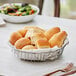 A round chrome wire basket filled with bread rolls on a table.