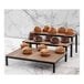 A table with three Abert Revolution wooden display risers holding muffins in a bakery.