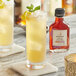 A glass of yellow liquid with ice and a mint leaf with a bottle of Runamok Aromatic Maple Bitters.