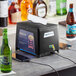 A black Powers Paper interfold napkin dispenser with a cell phone charging station on a counter next to a phone and bottles.