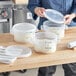 A person pouring white dough into a Vigor translucent plastic food storage container with a translucent lid.