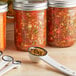 A white measuring spoon filled with Ball Fiesta Salsa Mix next to a jar of salsa.