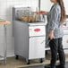 A woman standing in a professional kitchen using an Avantco natural gas floor fryer.