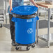 A woman in a blue uniform pushing a Lavex blue commercial recycling can with a blue lid and dolly.