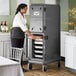 A woman in a white shirt and black pants standing in a professional kitchen putting food into a large black Cambro Pan Carrier.