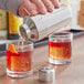 A hand pouring liquid from an Acopa stainless steel cobbler cocktail shaker into a glass.