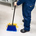 A warehouse broom with blue flagged bristles and a 48-inch handle being used to sweep debris on a tiled floor.