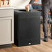 A man standing in front of a Rubbermaid black steel designer square trash can on a table in a coffee shop.